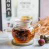 A glass tea pot with a blooming flowering tea next to some dried flowering teas