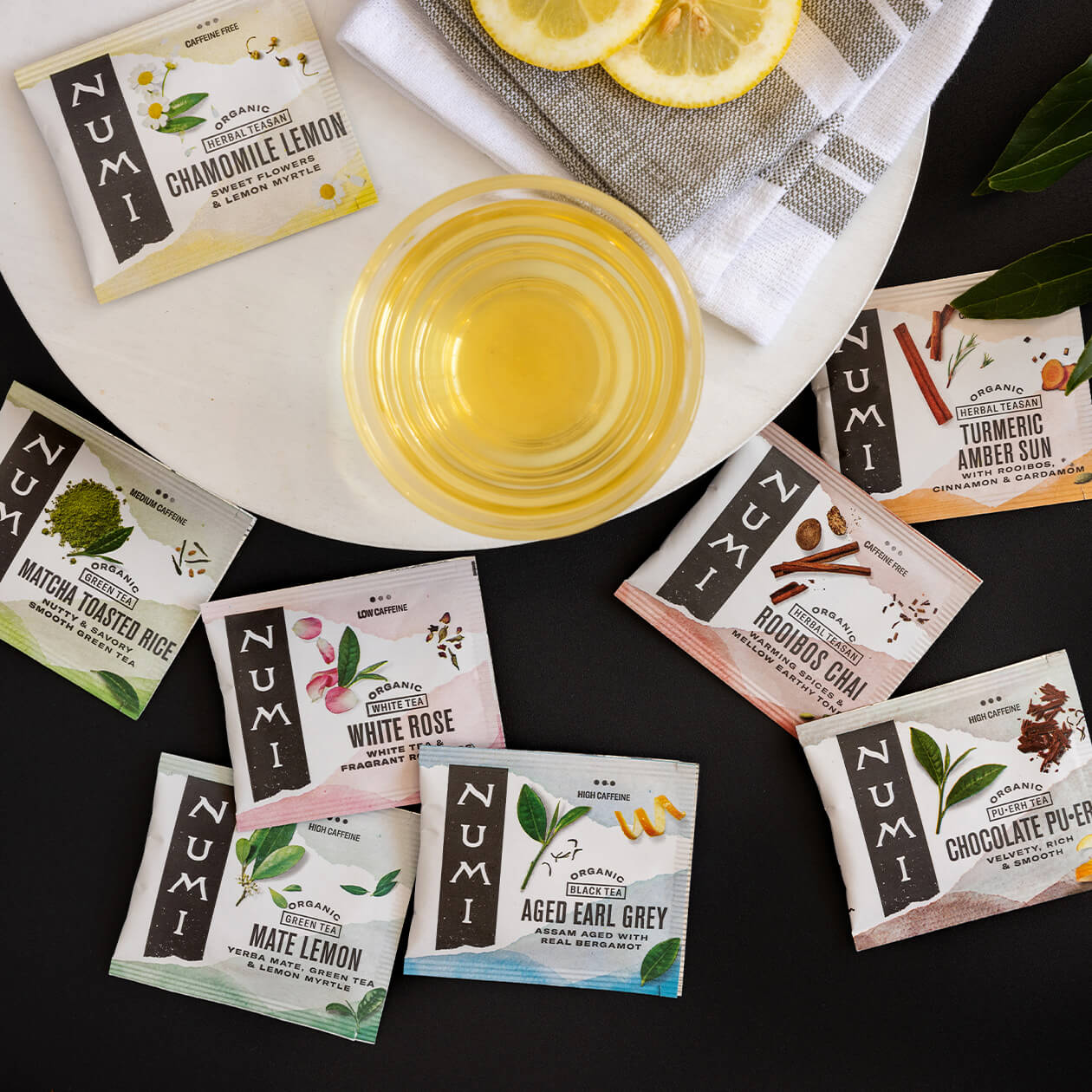 A bunch of Numi's Collection tea bags on a dark background with a cup of brewed tea