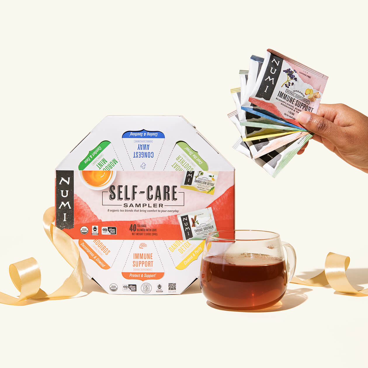 A Numi Self-care Sampler gift with a ribbon, a cup of brewed tea, and a person holding the variety of tea bags that come in this gift