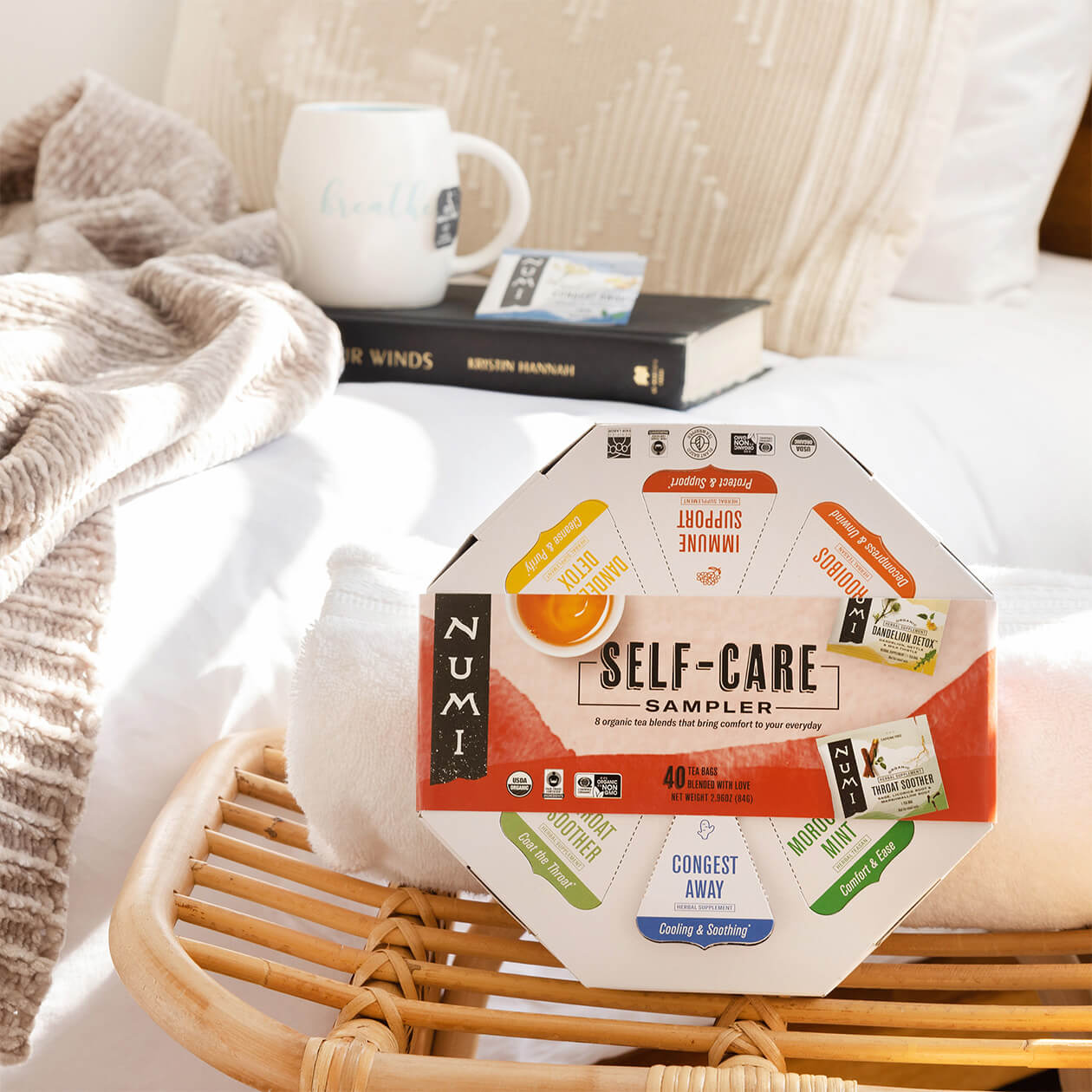 A Numi Self-care Sampler gift sitting on a bench next to a bed with a brewed cup of hot Congest Away tea