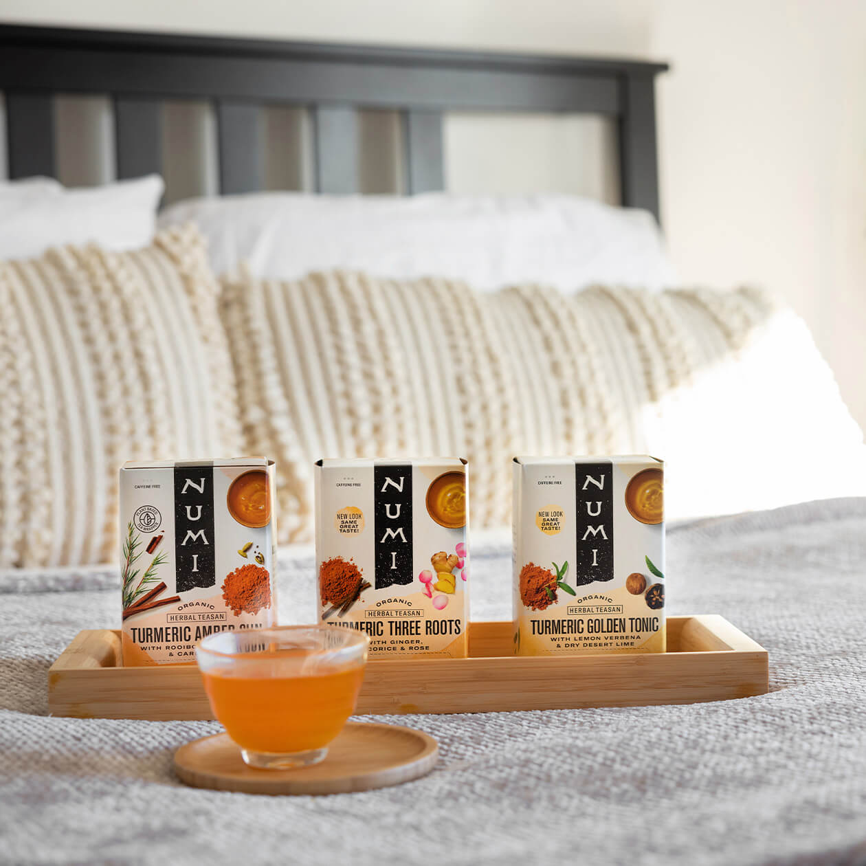Three boxes of Numi Turmeric Teas on a wooden tray on a bed with a hot cup of tea