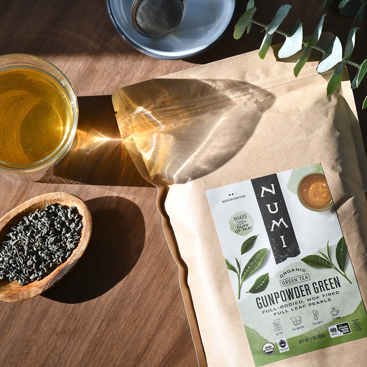 A bag of Gunpowder Green loose leaf tea sits on a wood table in the sunlight next to a brewed cup of loose leaf tea