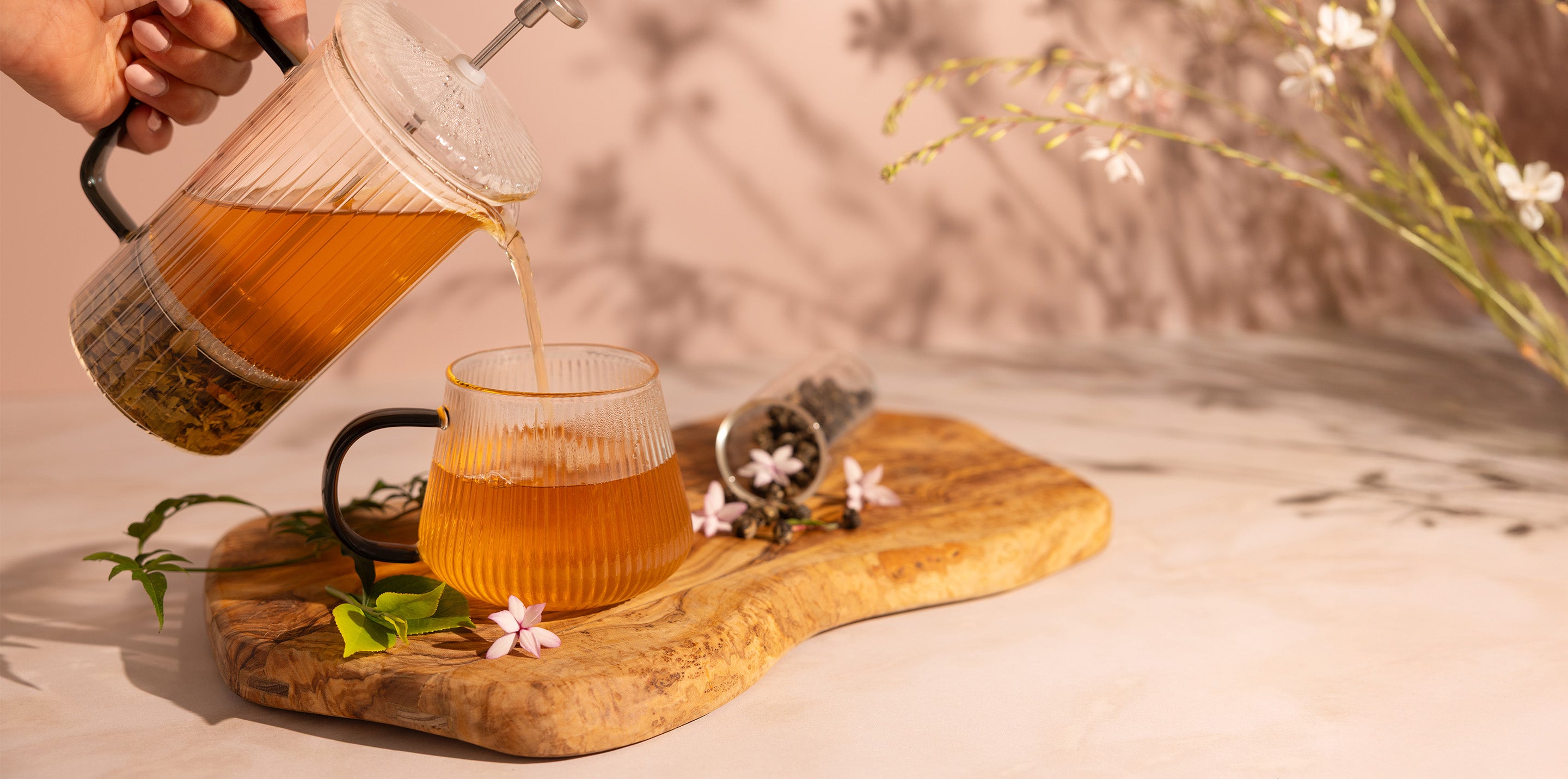 Tea being poured from a glass teapot into a cup on a wooden board with flowers.