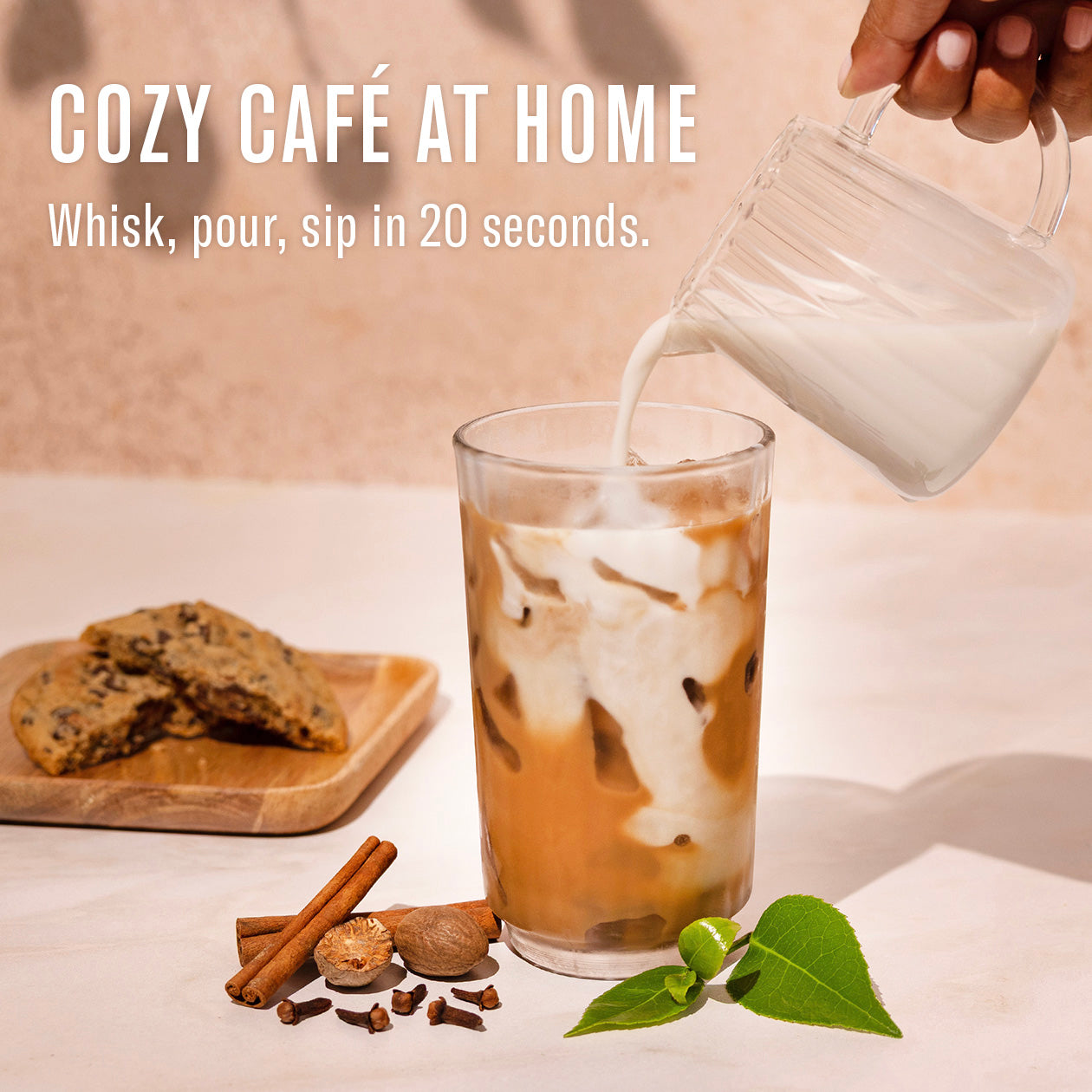 Glass of iced coffee with milk being poured, cookies and cinnamon sticks on a wooden board.