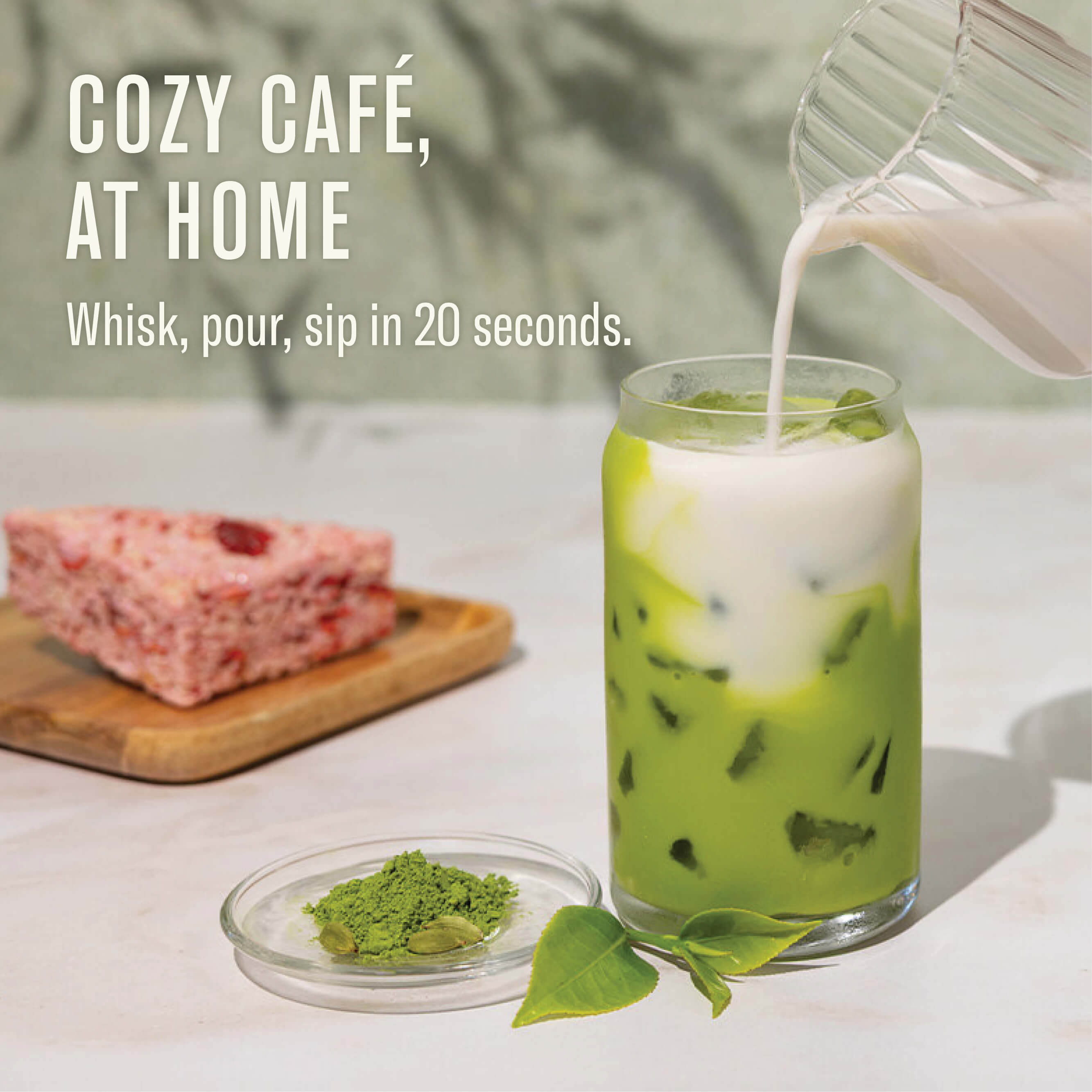 Green iced matcha drink being poured into a glass with a block of pink cake and matcha powder on a white surface.