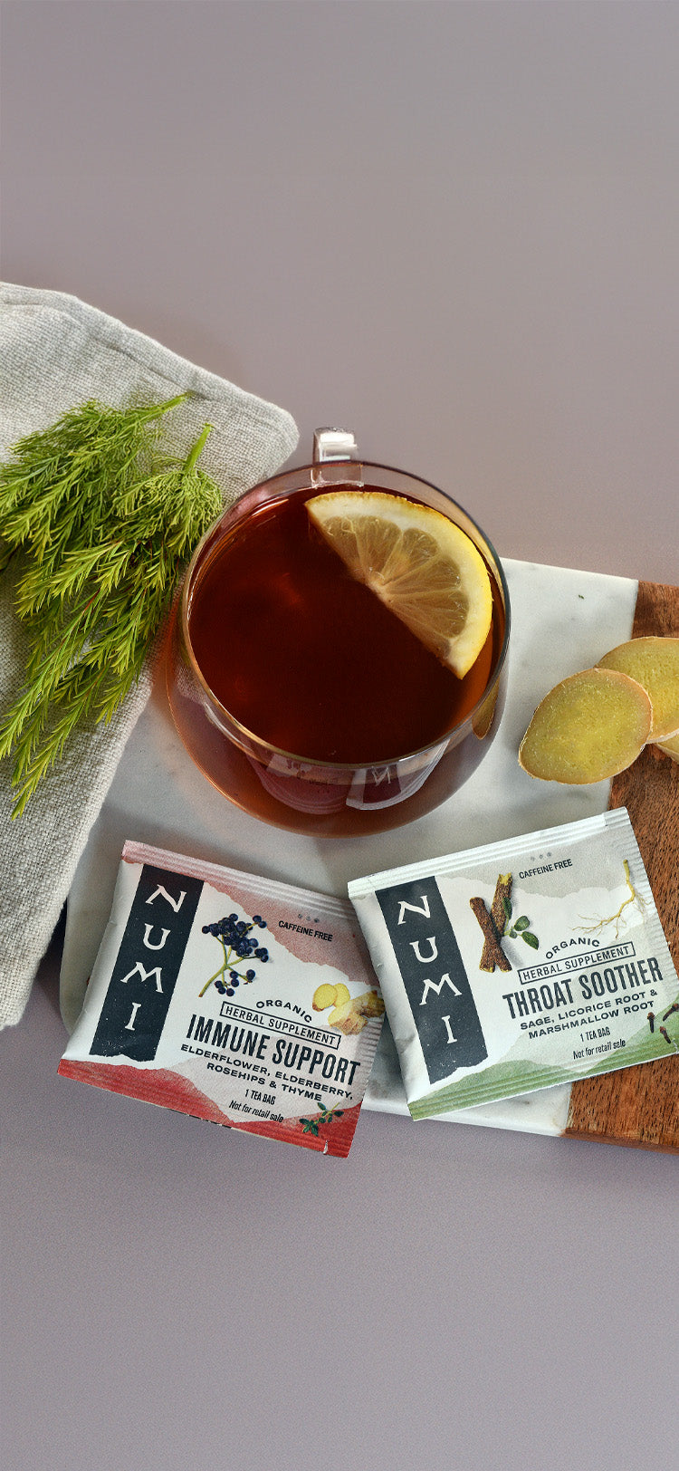 Two Numi tea bags on a gray surface with a cup of tea and garnishes.