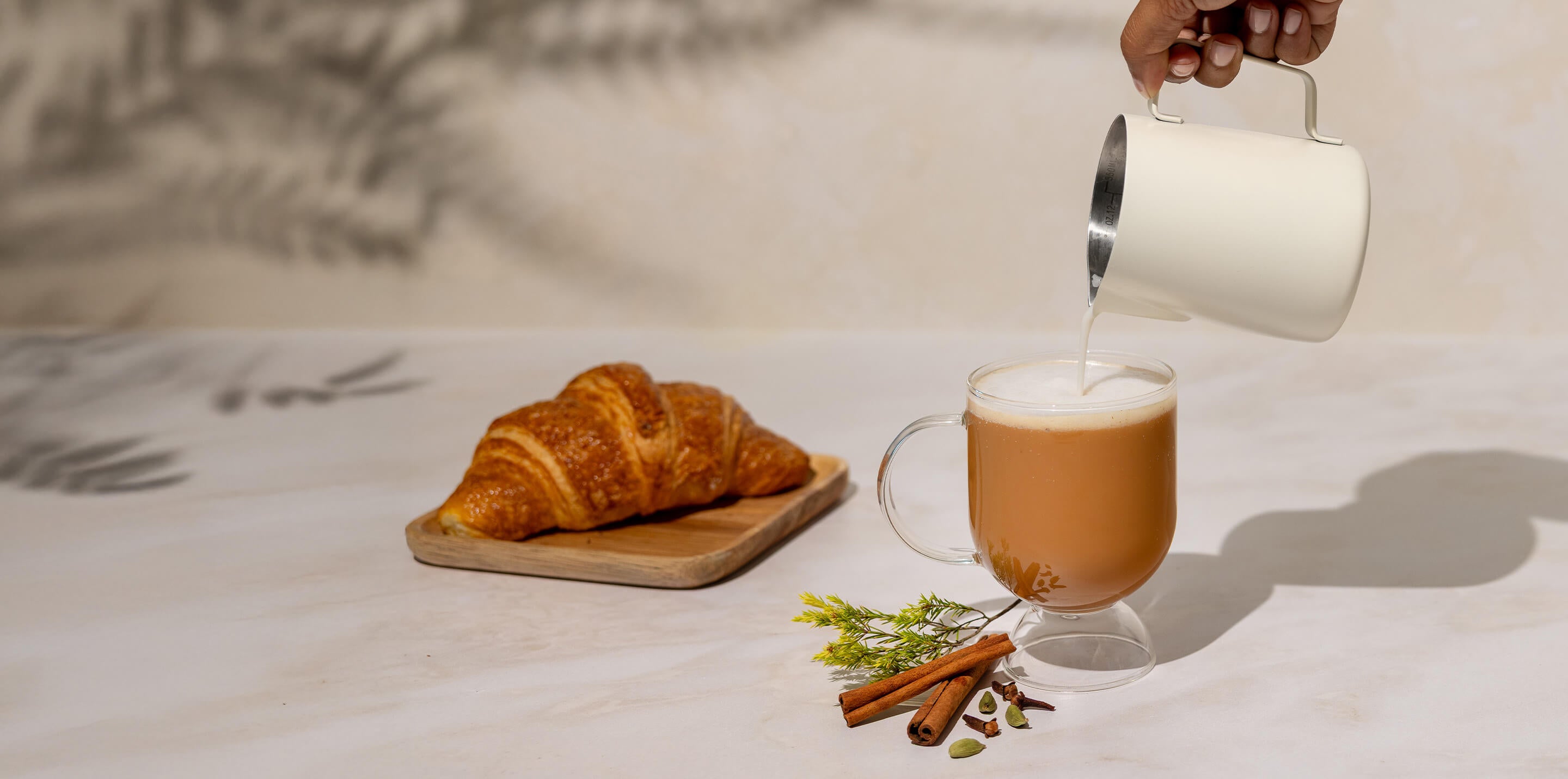 Person pouring a hot Latte into a glass mug with a croissant on a wooden board.