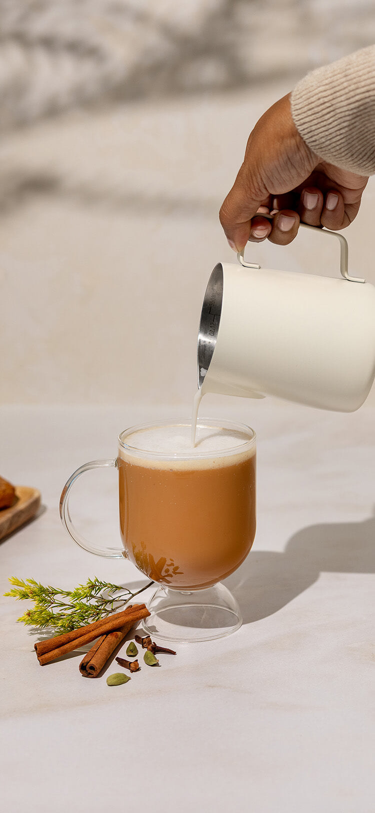 Person pouring a hot beverage into a glass mug with cinnamon sticks and a sprig of rosemary on a white surface.