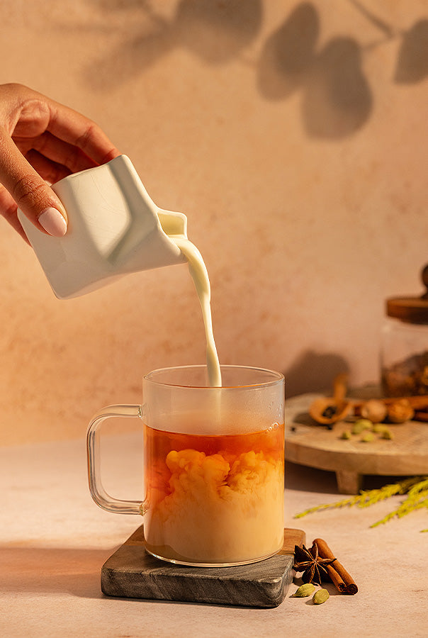 Person pouring milk into a glass of tea with a warm, homey background.