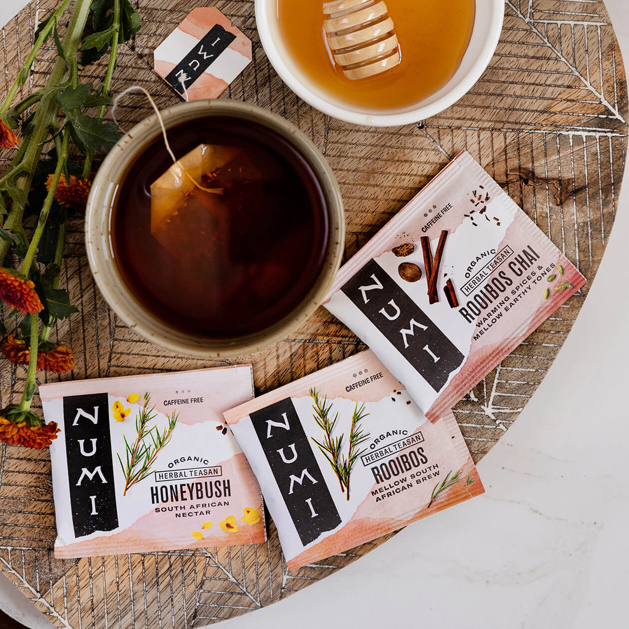 A brewing cup of tea, bowl of honey, flowers and Honeybush, Rooibos and Rooibos Chai tea bags on a wood tray