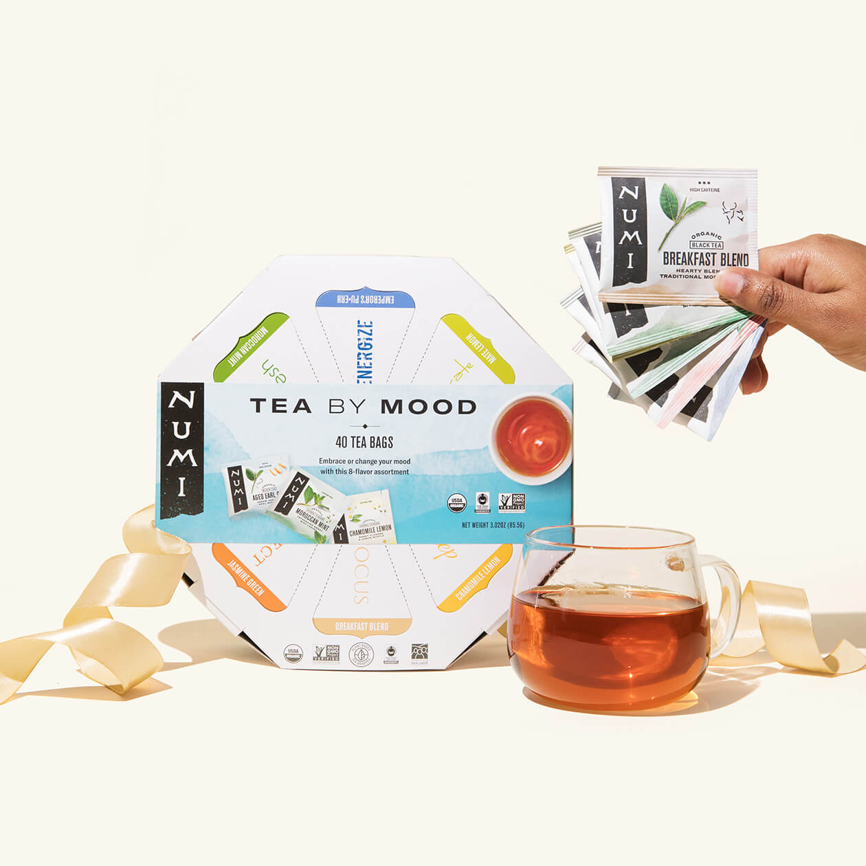 A Numi Tea By Mood gift with a ribbon, a cup of brewed tea, and a person holding the variety of tea bags that come in this gift